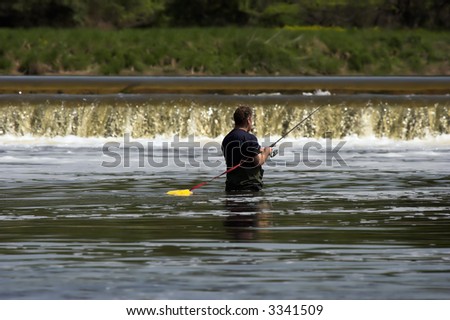 Man wading in a river and fly fishing with a low waterfall in the background.