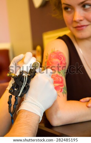tattooer showing process of making a tattoo on young beautiful hipster woman with red curly hair hand. Tattoo design in the form of rose. Focus on tattoo machine
