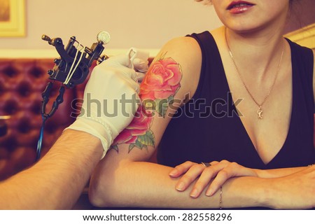 tattooer showing process of making a tattoo on arm of young beautiful hipster woman with red curly hair. Tattoo design in the form of roses. Vintage style picture