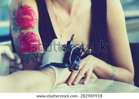 tattooer showing process of making a tattoo on young beautiful hipster woman arm. Tattoo design in the form of rose. Vintage picture