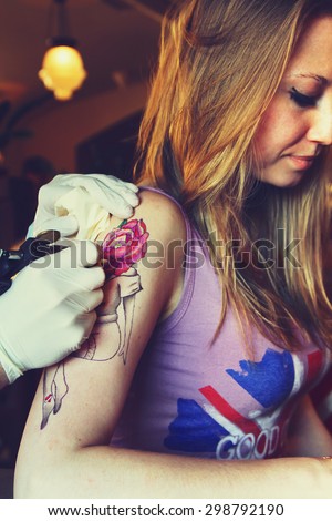 tattooer showing process of making a tattoo on young beautiful hipster woman with blonde  hair arm. Tattoo design in the form pin-up girl with flower head. Vintage picture