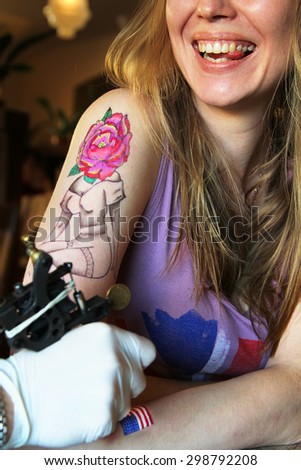 tattooer showing process of making a tattoo on young beautiful hipster woman with blonde hair hand. Tattoo design in the form pin-up girl with flower head