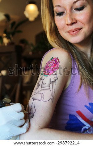 tattooer showing process of making a tattoo on young beautiful hipster woman with blonde hair hand. Tattoo design in the form pin-up girl with flower head