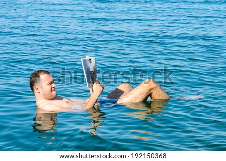 DEAD SEA, JORDAN - MAY 1, 2014: Unidentified man reads Ricky Martin book laying on the surface of  the Dead Sea water, Jordan. Dead Sea water is used for medical purposes for people with skin problems
