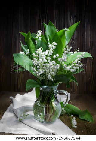 Flowers of Lilies of the valley in a glass jug on the wooden background. Light brush technique.