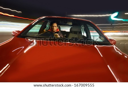 Looking through the windscreen at a man driving a red sports car at night.