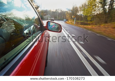 A man driving a red car towards a Speed camera on a country lane.