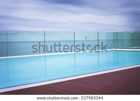 Deserted cool blue inviting swimming pool on a paved patio overlooking the ocean surrounded by a wire mesh fence, , idyllic summer background