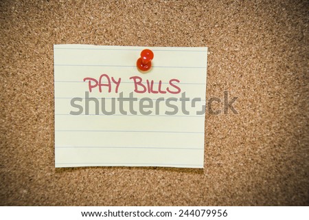 Yellow note attached with pushpin to a bulletin board contain a hand written reminder to pay bills