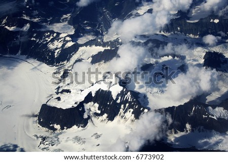 black mountains viewed from above covered with white glacier with clouds floating above it.