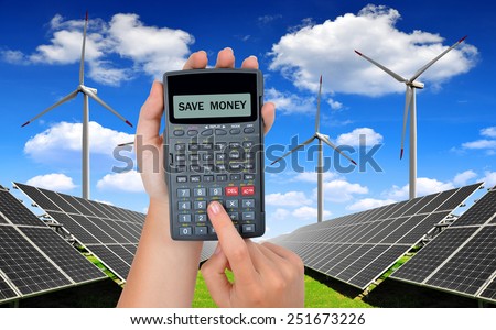 Hand with calculator. In the background solar energy panels and wind turbines.Concept of saving money.