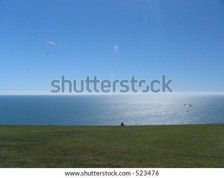 A lone person sat in a green field looking out to sea with hang gliders.