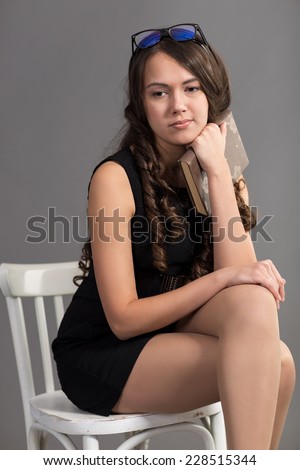brunette with long hair in black dress sits on white chair with book