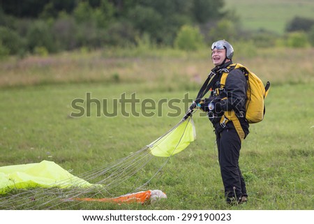 Joyful man in a black suit with a yellow parachute standing on the green field after the jump