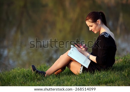 Happy woman lying on the grass and writing in a notebook with unfocused lake in the background