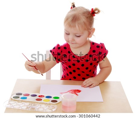 Pretty blonde little girl in a red polka-dot dress sitting at the table. She paints watercolors on the white paper-Isolated on white background