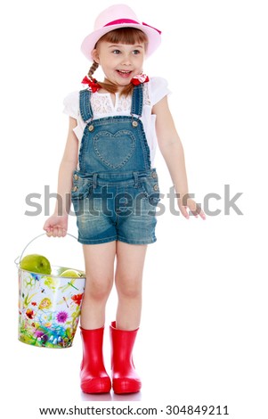 The little blonde girl in denim overalls holding a bucket of green apples. Head girls pink hat,braided pigtails red bows-Isolated on white background