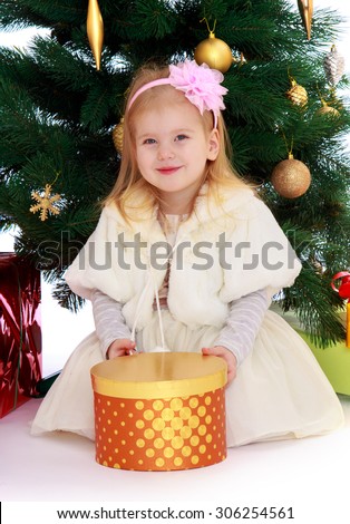 Beautiful little blonde girl in a Princess dress on the Christmas tree. Before the girl stands round box with a gift-Isolated on white background