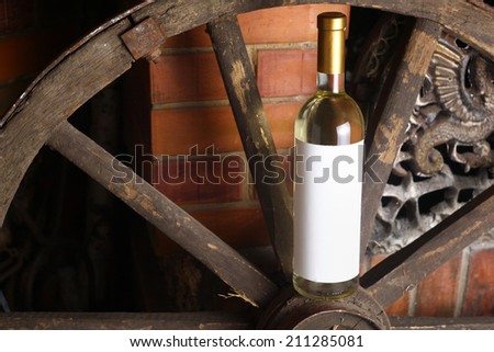 Bottle of white wine with blank label standing on an old wooden wheel near a fireplace