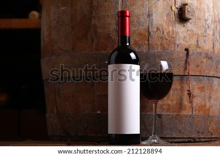 Bottle of red wine with a blank label and glass standing on cellar floor with a wooden wine barrel in the background