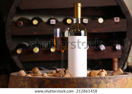 Bottle of white wine with blank label template and glass standing in a wine cellar on a wood barrel