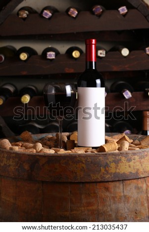 Bottle of red wine with blank label template and glass standing in a wine cellar on a wood barrel