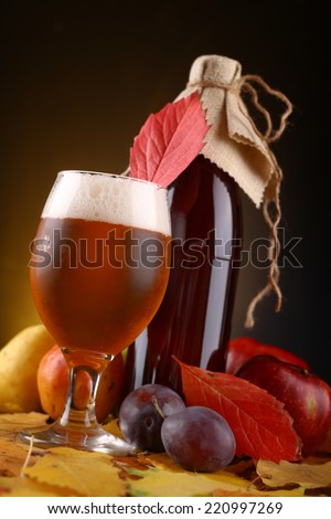 A glass and a bottle of beer standing over yellow leaves with an assortment of fruits over a dark warm background