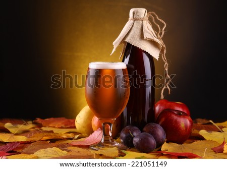 A glass and a bottle of beer standing over yellow leaves with an assortment of fruits over a dark warm background