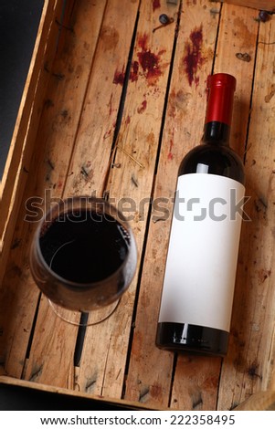 Bottle of red wine with a blank label and a full glass in a used wooden crate with grape juice marks