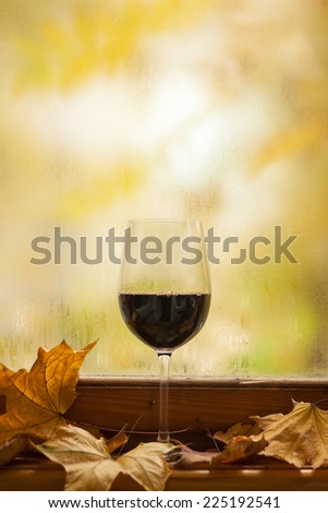 Glass of red wine standing on windowsill with autumn leaves and a fogged window in the background