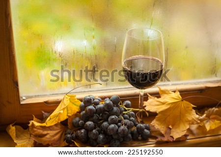 Glass of red wine standing on windowsill with autumn leaves and grapes with a fogged window in the background