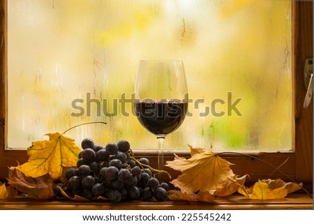 Glass of red wine standing on windowsill with autumn leaves and grapes with a fogged window in the background