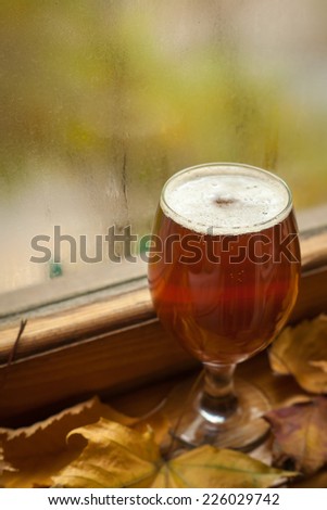 Glass of amber beer standing on windowsill with autumn leaves and a fogged window in the background