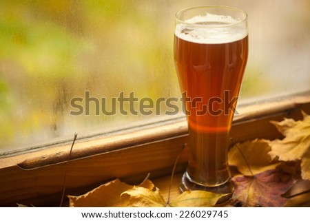 Tall glass of amber beer standing on windowsill with autumn leaves and a fogged window in the background