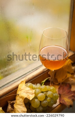 Glass of white wine standing on windowsill with autumn leaves and white grapes with a fogged window in the background