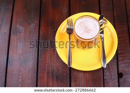 A classic mug full of light beer standing on a plate with knife and fork on a wooden table