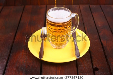 A classic mug full of light beer standing on a plate with knife and fork on a wooden table