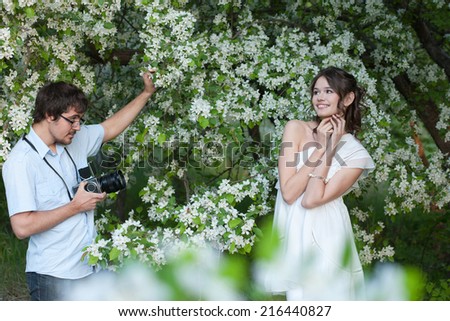 The man in a blue shirt photographs the young girl in a white dress under branches of a blossoming apple-tree