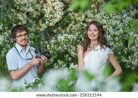 The man in a blue shirt photographs the young girl in a white dress under branches of a blossoming apple-tree