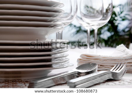 Setting the table with wine glasses, dishes, napkins, forks and spoons over a embroidered linen tablecloth. Window at the background.