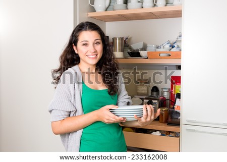 Happy young woman taking out dishware of the cupboard to set the table