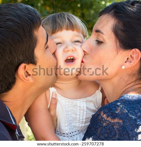 Mum and dad kissing tight baby face outdoors in a warm day