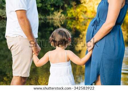 Pregnant family holding hands on the shores of a river. Back view. Concept of union, support and future.