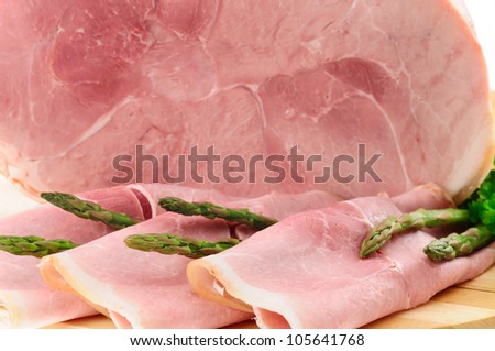 Slices of York ham with green asparagus tips and whole ham cross-section as background. Selective focus.