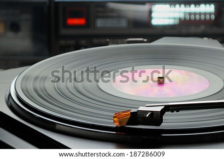 Retro record player turntable with vinyl record (LP) with lit sound level meter meter in background and other electronic equipment. Selective focus.