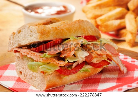 Bacon, lettuce and tomato (BLT) sandwich from freshly cut baguette on rustic wooden bread board. French fries and tomato ketchup on background.