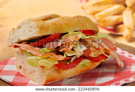 Bacon, lettuce and tomato (BLT) sandwich from freshly cut baguette on rustic wooden bread board with French fries in background.