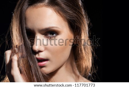 Close-up portrait of a pretty brunette girl on the black background.Shadows.Make-up.\
Model  hand touches the hair, her face half covered by hair.Shadow theater, mysticism