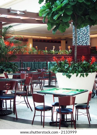 shot of some tables and chairs in a food court at a mall. a lots of plants are decorating the place.