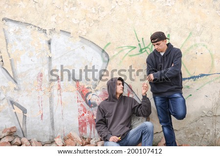 two street hooligans or rappers standing against a graffiti painted wall and sharing a cigarette